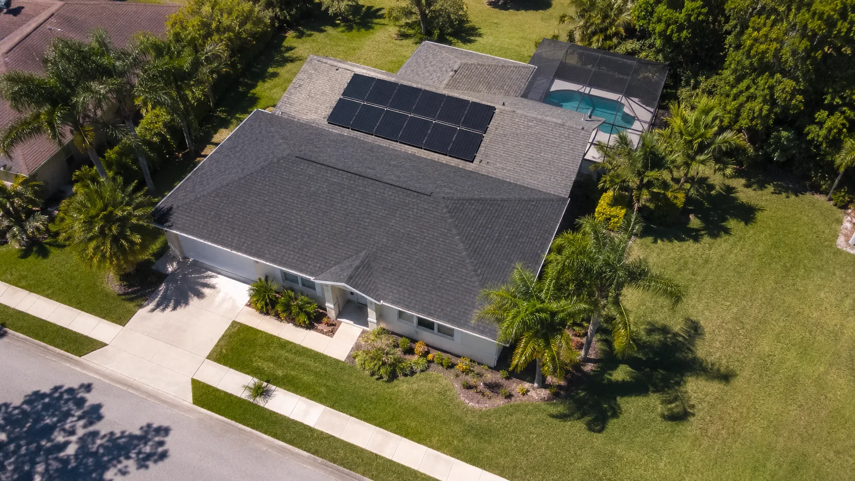 Aerial view of a single-story Florida home with a solar array on one roof plane, a screened pool enclosure behind it and palms around the lot